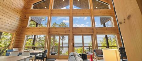 A spacious room with a high ceiling and large windows offering a view of trees and a body of water.