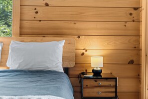 Een houten bed met een wit kussen en een blauwe deken, een bedlampje en een klein houten nachtkastje met lades.