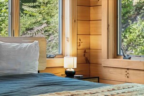 Ein Schlafzimmer mit einem Bett, einem Nachttisch, einer Lampe und einem Fenster mit Blick auf Grünflächen.