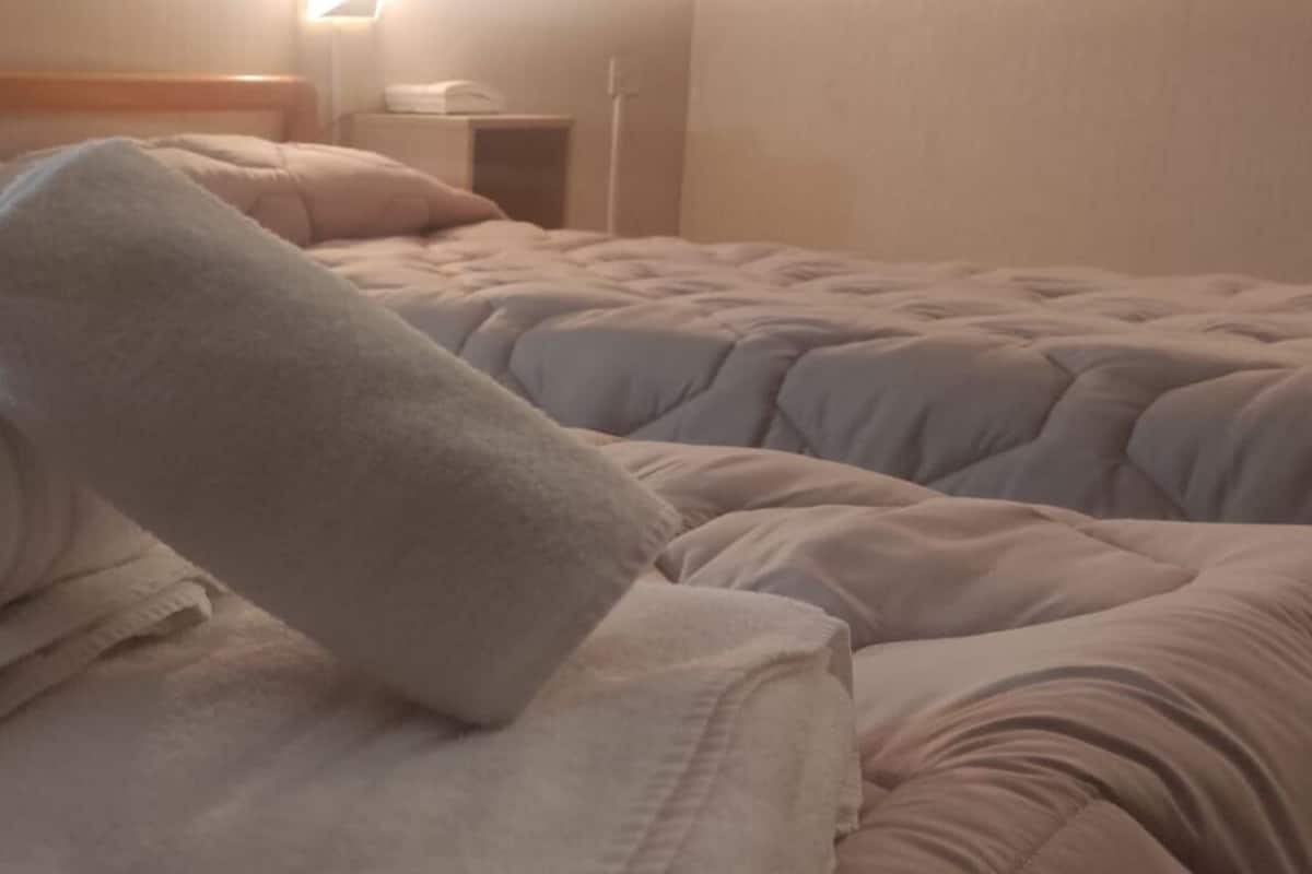 A hotel room with two beds, each with a white duvet and pillows. There is a nightstand with a lamp and a small table.