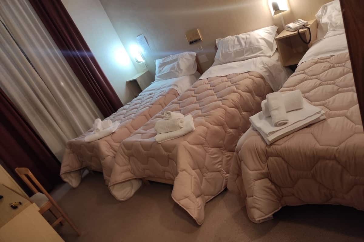 A hotel room with two beds, each with a beige quilted bedspread and white pillows. There are towels on the beds, and a bedside table with a lamp and a phone.