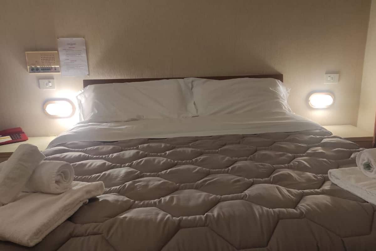 A neatly made bed with a beige quilt, two bedside lamps, a control panel, and a set of towels on the side.