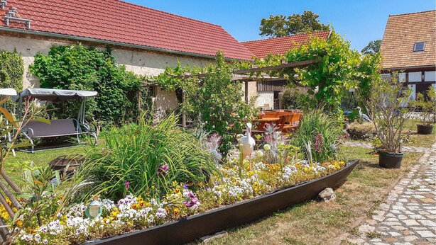 Un jardín con diversas plantas y flores, una banca de madera y una pérgola.