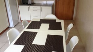 Une petite cuisine avec des meubles blancs, un four à micro-ondes et une porte en bois. On y trouve une table avec quatre chaises et un set de table à carreaux.