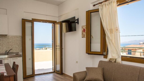 A living room with a sofa, a television mounted on the wall, and a view of the sea through a sliding glass door. - Apartment for 3 pax | Bellino Apartments (Chersonissos)