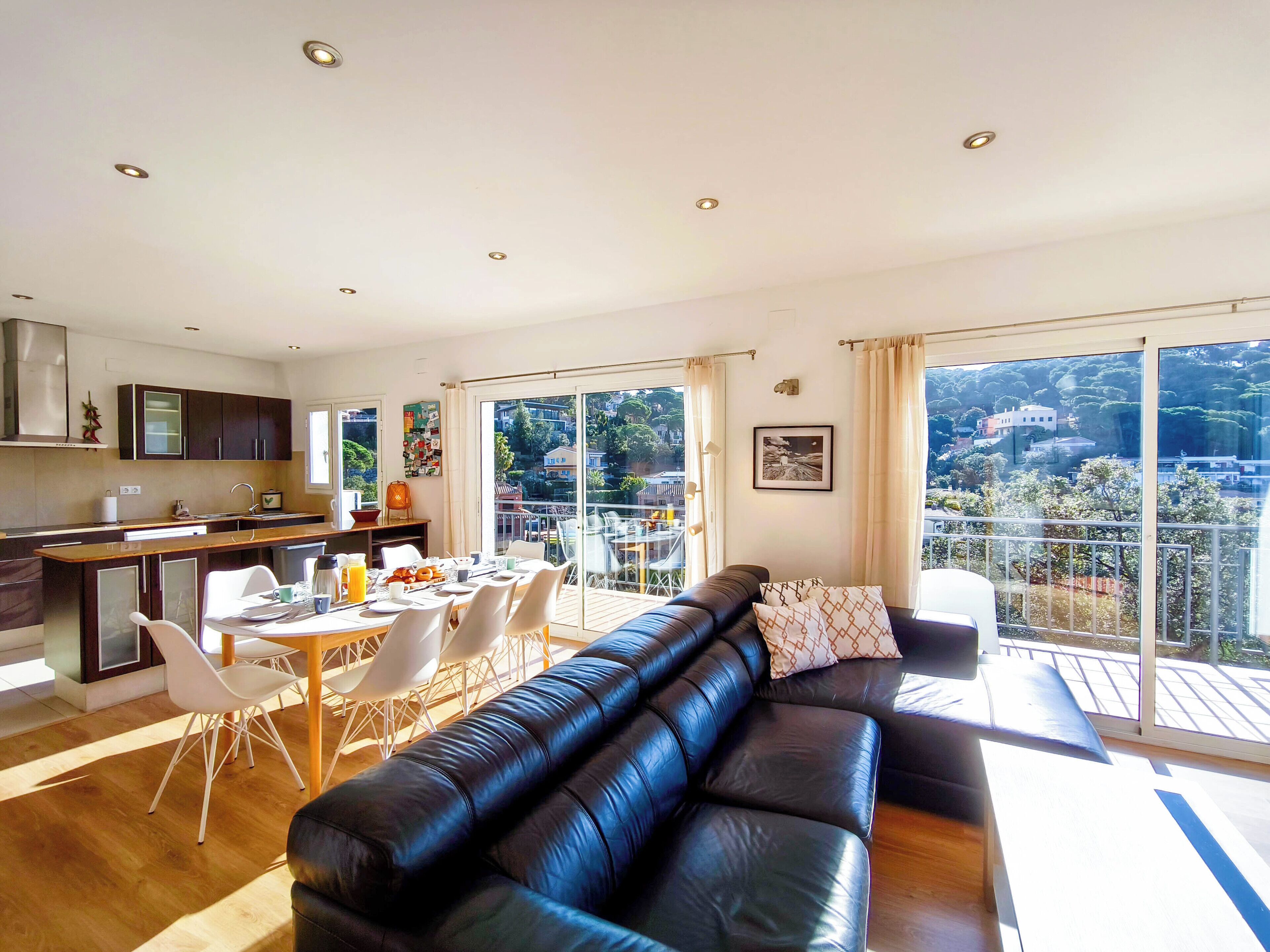 A modern living room with a black leather sofa, dining area, kitchen, and large windows offering a city view.
