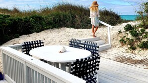 A deck with a round table and chairs, overlooking a beach with sand and vegetation. - Beach Front Villa (Abaco)