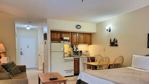 Una habitación compacta con cama, una mesita, una silla, kitchenette y una puerta.