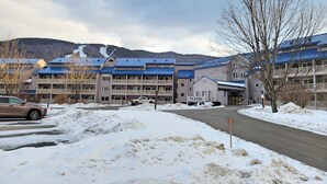 Un hotel de varios pisos con techo azul, un estacionamiento nevado y montañas al fondo.
