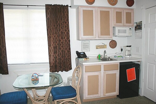 A compact kitchenette with a microwave, coffee maker, and a small sink. There is a dining table with chairs and a decorative item on the wall.