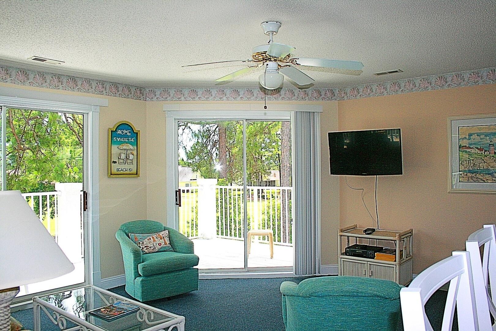 A room with a ceiling fan, a television, a green armchair, a glass coffee table, and a view of the outdoors through large sliding glass doors.