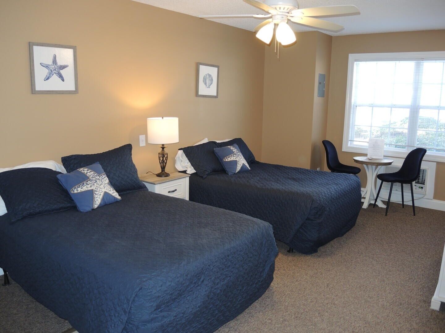 A hotel room with two beds, a desk, a chair, and a ceiling fan.