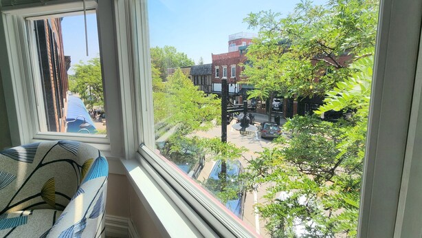 A view from a window with a patterned curtain, overlooking a street with buildings, trees, and parked cars. - Summer Fling 2nd Floor Downtown Suite  (South Haven)