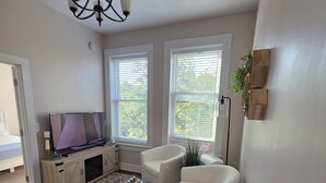 A living room with a sofa, armchairs, a coffee table, and a TV. There are windows with blinds, a chandelier, and a plant on the wall. - Summer Fling 2nd Floor Downtown Suite  (South Haven)