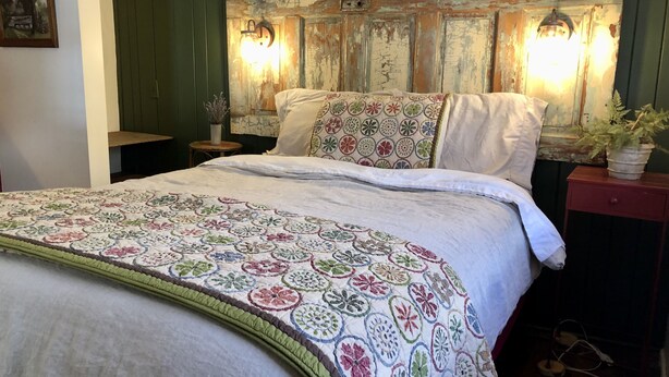 A bed with a floral-patterned quilt, a rustic headboard with wall-mounted lights, and a bedside table with a potted plant. - Adirondack Retreat - Hot Tub & Lake (Keeseville)