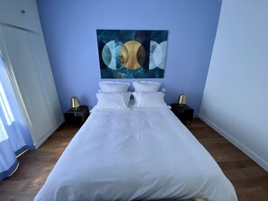 A neatly made bed with white linens in a room with light blue walls and a large abstract painting above it.