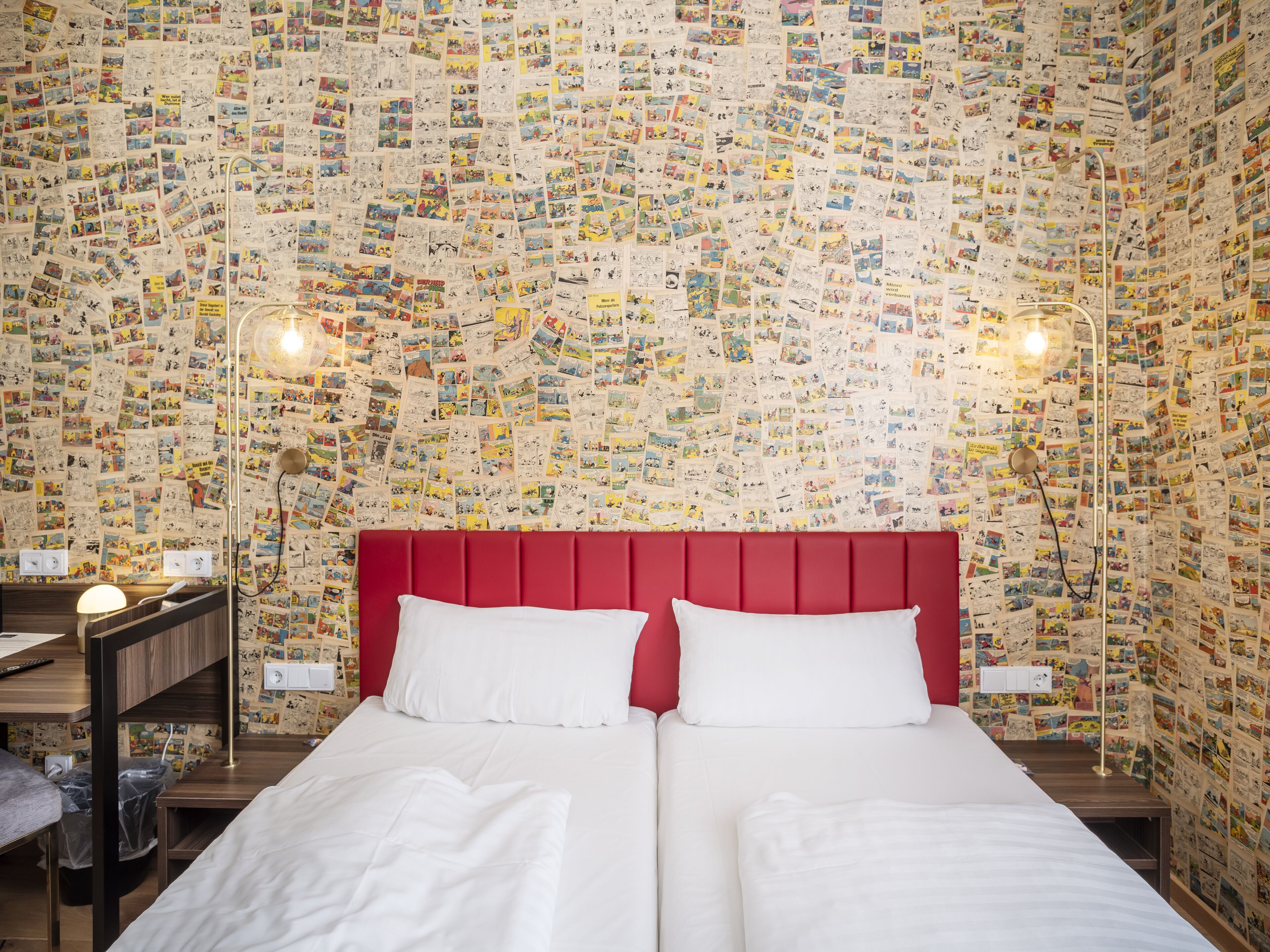 A hotel room with a red headboard, two white pillows, a wooden nightstand, a lamp, and a wall covered with postcards.