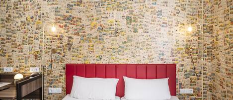 A hotel room with a red headboard, two white pillows, a wooden nightstand, a lamp, and a wall covered with postcards.