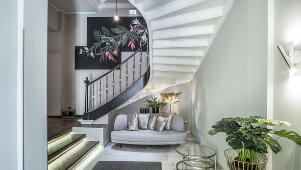 A modern interior with a curved staircase, a grey sofa with patterned cushions, a glass coffee table, and a large potted plant. - acor Boutique Hotel (Heidelberg)