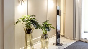 A modern hallway with a light-colored floor, two potted plants in gold wire stands, a wall-mounted light, and a door with a glass panel. - acor Boutique Hotel (Heidelberg)