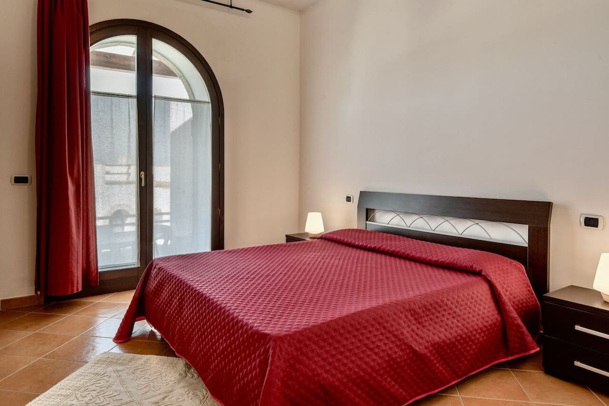 Quarto com cama grande, mesa de cabeceira com abajur, janela com cortinas e vista para o exterior.