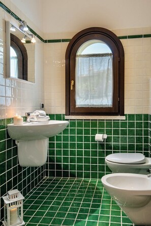 A bathroom with a green-tiled floor, a white sink, a white toilet, and a window with a curtain.