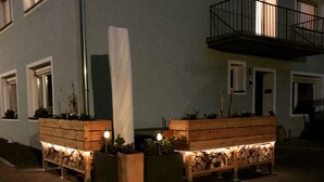 Vista nocturna de un edificio con balcón, macetas de madera con plantas y un sendero iluminado.
