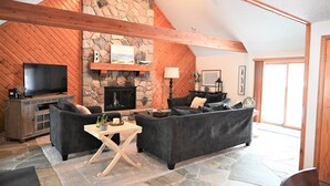 A cozy living room with a stone fireplace, wooden beams, and a flat-screen TV. - Lake Front, Lake Charlevoix, Large Park like setting. (Boyne city)