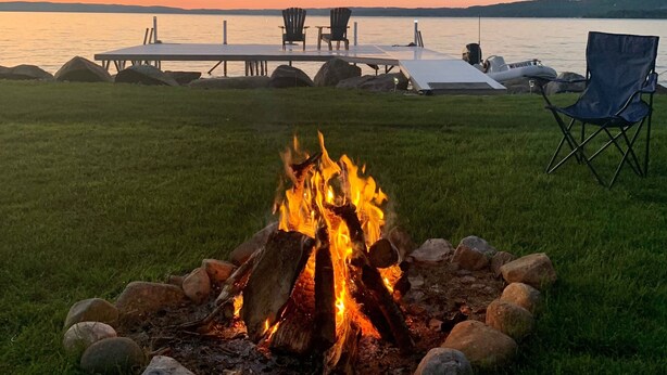 A fire pit with rocks and logs in a grassy area, a dock with chairs, and a calm lake at sunset. - Lake Front, Lake Charlevoix, Large Park like setting. (Boyne city)