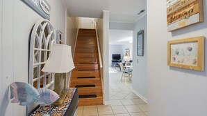 A staircase leading to a living area with a television, a dining table, and chairs. There are wall decorations and a lamp on a console table.
