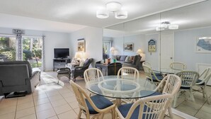 A spacious living area with a glass table surrounded by wicker chairs, a grey sofa, and a television.