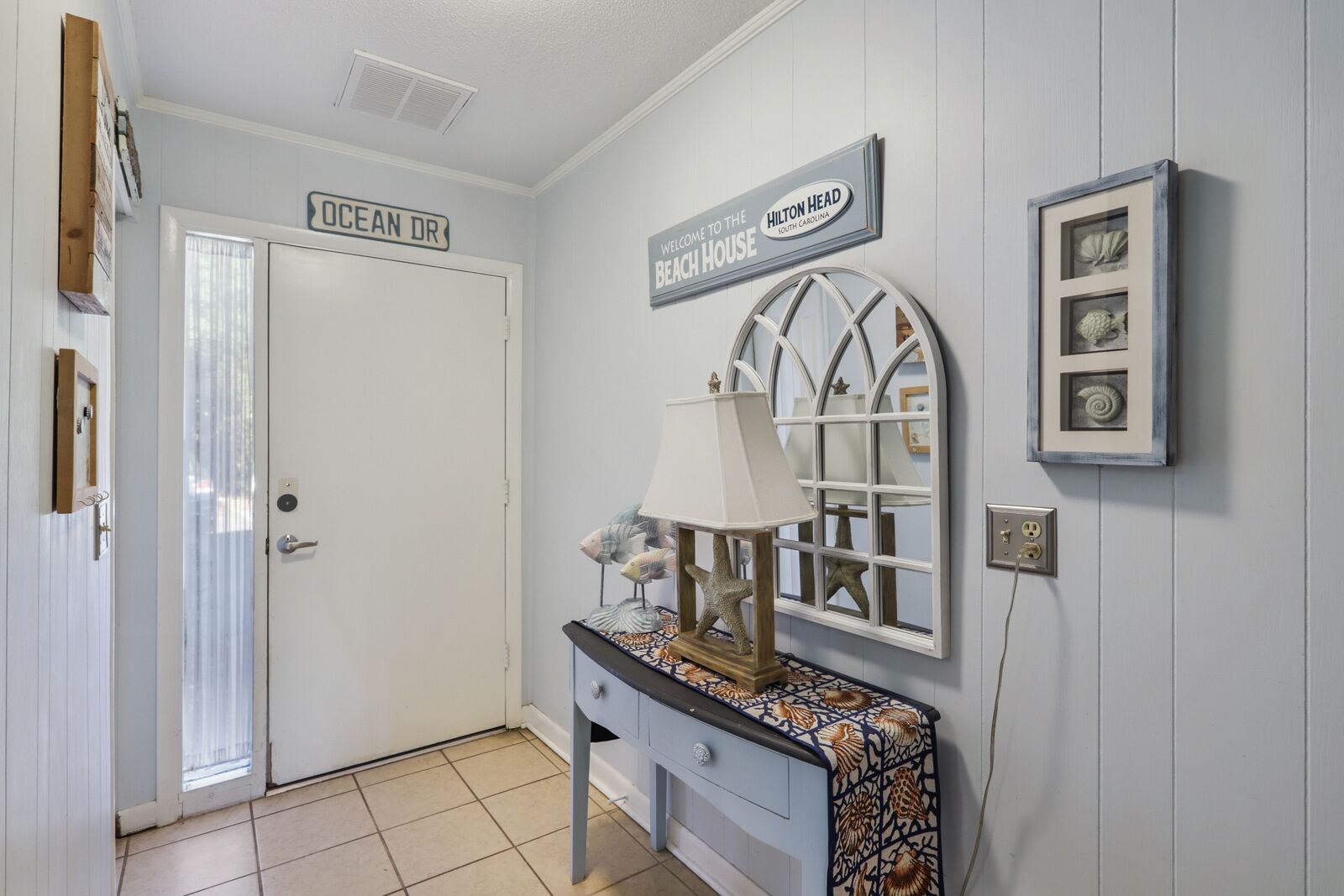 Un corridoio con una porta etichettata 'OCEAN DR', un cartello di benvenuto per la casa sulla spiaggia, una lampada decorata con stelle marine e un quadro appeso al muro.