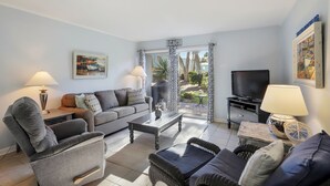 A living room with a sofa, armchair, coffee table, TV, and a view of a garden through a glass door.
