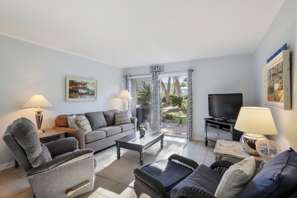 A living room with a sofa, armchair, coffee table, TV, and a view of a garden through a glass door.
