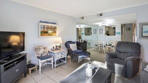 A living room with a television, two chairs, a dining table, and a kitchen area visible through a glass door.