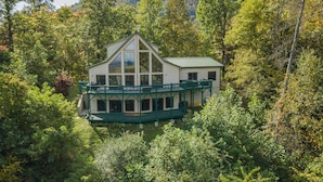 A two-story house with a green balcony, surrounded by dense forest. - Hummingbirds' Mountain Retreat | Hot Tub, Game Room, Home Office & Mountain Views! (Old Fort)