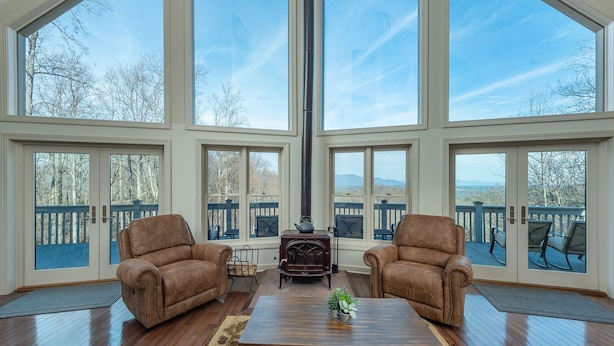 A spacious room with large windows, two leather armchairs, a wooden coffee table, and a wood-burning stove. - Hummingbirds' Mountain Retreat | Hot Tub, Game Room, Home Office & Mountain Views! (Old Fort)