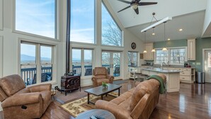 A spacious living room with large windows, a ceiling fan, and a kitchen area with a view. - Hummingbirds' Mountain Retreat | Hot Tub, Game Room, Home Office & Mountain Views! (Old Fort)