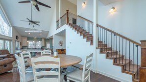 A spacious living area with a wooden dining table, white chairs, a staircase with a wooden railing, and a ceiling fan. - Hummingbirds' Mountain Retreat | Hot Tub, Game Room, Home Office & Mountain Views! (Old Fort)