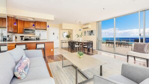 A modern living room with a sofa, coffee table, and dining area. A kitchen with wooden cabinets and appliances is visible. Large windows offer a view of the sky and clouds. - Ocean-Marina Views PH at Ilikai Hotel by the Beach w/Full Kitchen & Free WiFi (Honolulu)