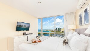 A modern hotel room with a large bed, a TV on a white dresser, a balcony with a city view, and a large window with curtains. - Ocean-Marina Views PH at Ilikai Hotel by the Beach w/Full Kitchen & Free WiFi (Honolulu)