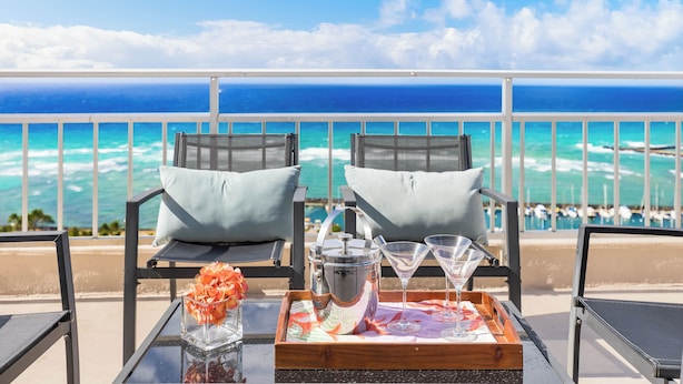 A balcony with two chairs, a tray with a drink pitcher, martini glasses, and a vase of flowers. - Ocean-Marina Views PH at Ilikai Hotel by the Beach w/Full Kitchen & Free WiFi (Honolulu)