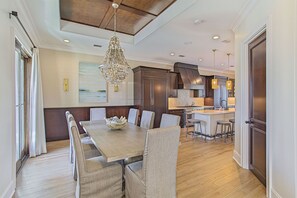 A dining area with a long wooden table, cushioned chairs, a chandelier, and a kitchen in the background.