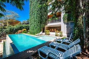 Una piscina con sillas de descanso, rodeada de vegetación y un edificio con balcones.