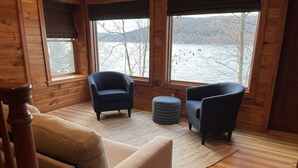 Ein gemütliches Blockhaus-Interieur mit einer Couch, zwei Sesseln, einem runden Hocker und großen Fenstern mit Blick auf einen See und Berge.