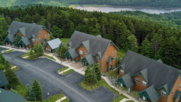 A cluster of log cabin-style buildings with steep roofs, surrounded by lush greenery and a lake in the background. - Camp Four 23: Pet Friendly,  Slope side, Gas Fireplace. (Snowshoe)