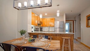 A modern kitchen with wooden cabinets, stainless steel appliances, and a dining area with a wooden table set for four. - Camp Four 23: Pet Friendly,  Slope side, Gas Fireplace. (Snowshoe)