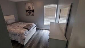 Un dormitorio con cama, mesita de noche, cómoda, ventana con persianas y una obra de arte colgada en la pared.