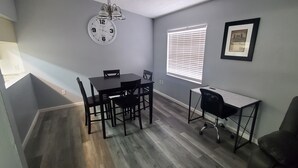 Un área de comedor con una mesa y sillas, un reloj, una ventana con persianas y un cuadro en la pared.