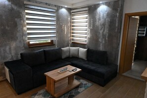 A modern living room with a sectional sofa, a wooden coffee table, and large windows with blinds.